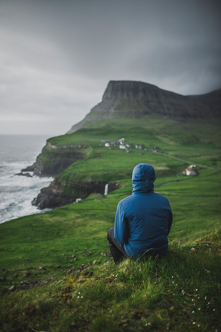 Mann sitzt mit Blick auf Klippenküste und Ozean, Gasadalur-Dorf, Färöer, Dänemark