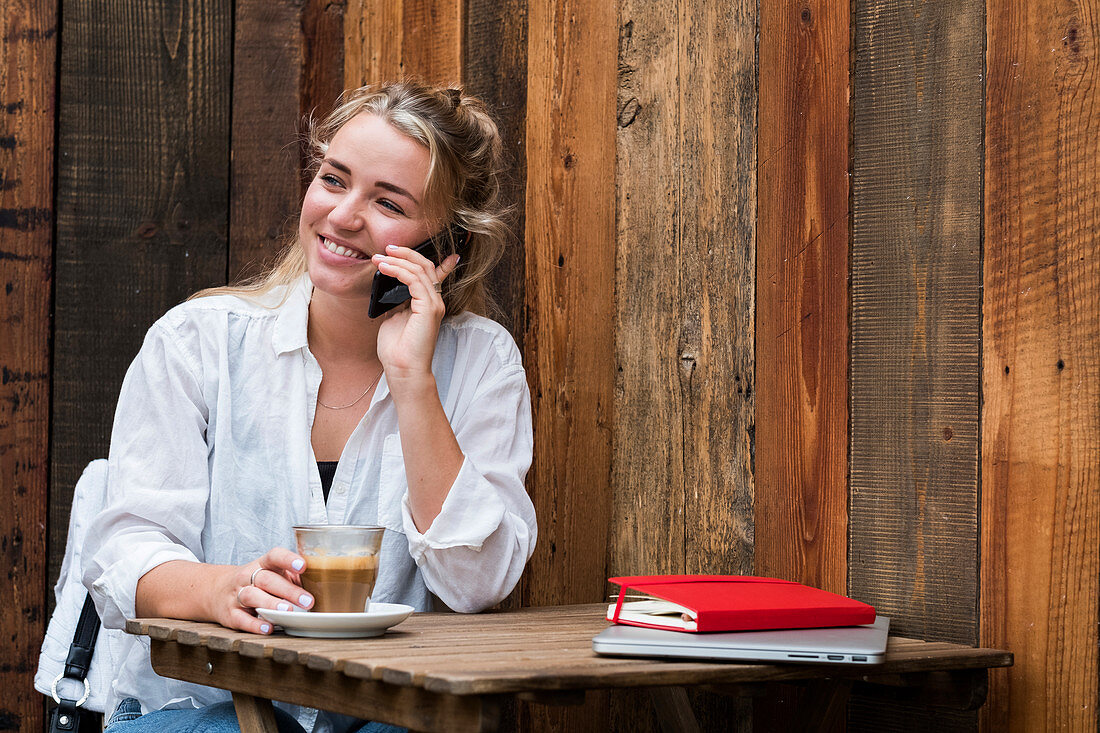 Junge blonde Frau, sitzt allein in einem Café, benutzt Smartphone, arbeitet aus der Ferne