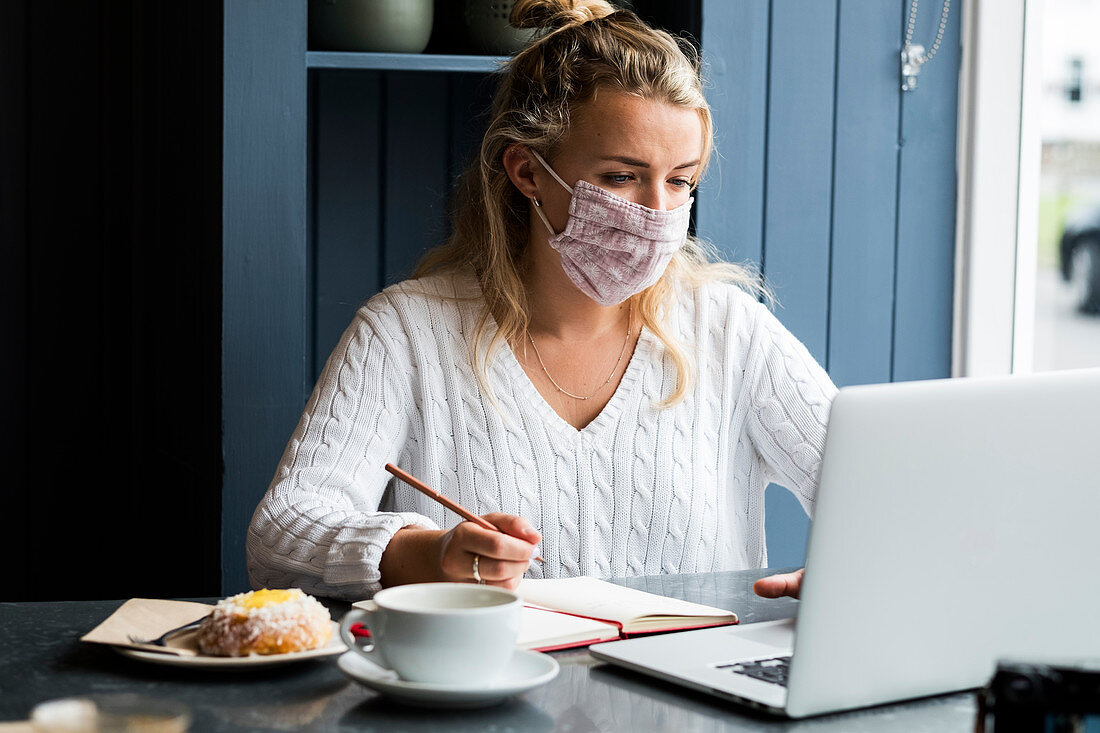Young blond woman wearing face mask sitting alone at a cafe table with a laptop computer, writing in note book, working remotely.