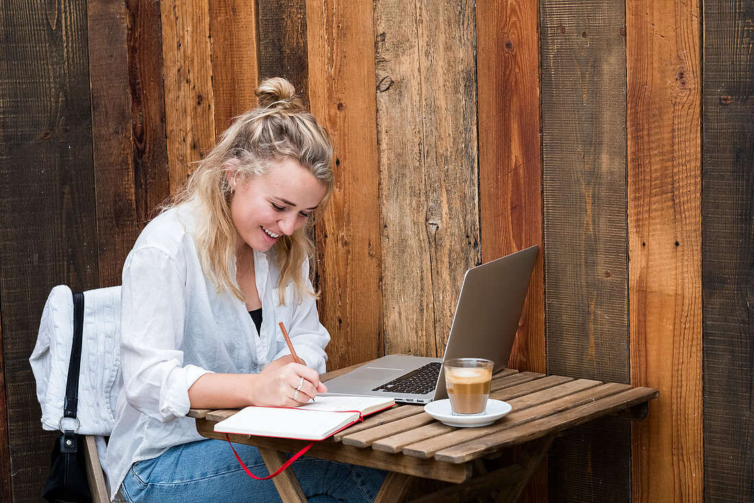Young blond woman sitting alone at a cafe table with a laptop computer, writing in note book, working remotely.
