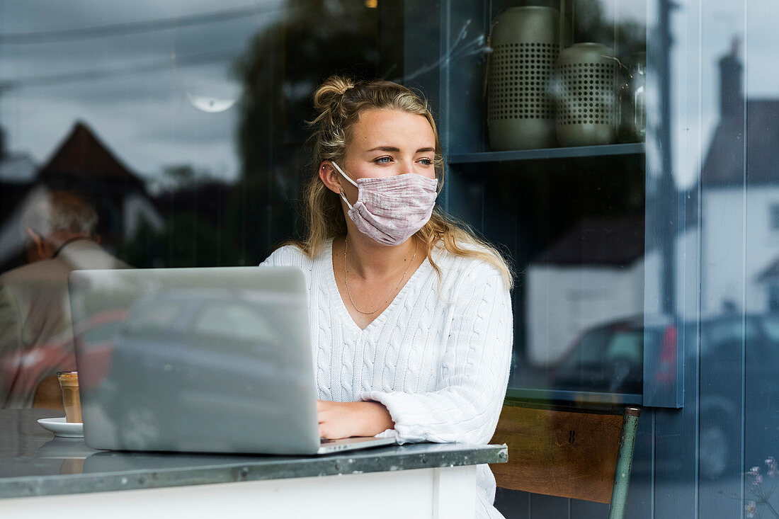 Junge blonde Frau mit Gesichtsmaske sitzt allein an einem Cafétisch mit einem Laptop, schreibt in Notizbuch, arbeitet aus der Ferne