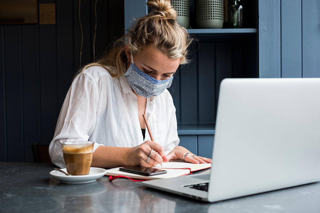 Junge blonde Frau mit Gesichtsmaske sitzt allein an einem Cafétisch mit einem Laptop, schreibt in Notizbuch, arbeitet aus der Ferne