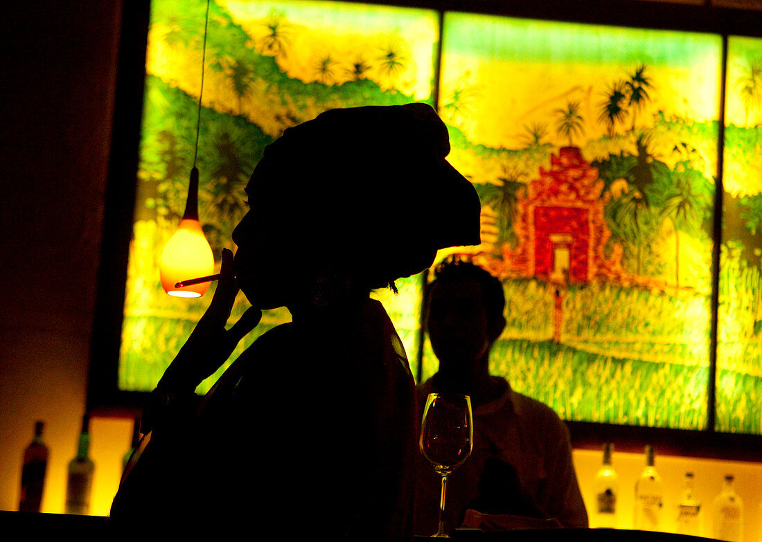 Eine Frau mit Kopftuch, die eine Zigarette raucht, Silhouette gegen ein beleuchtetes Dschungelszenenbild an einer Bar. In Bali, Indonesien, Asien