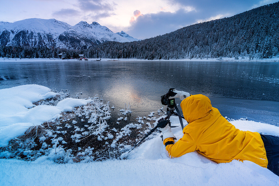 Photographer taking pictures of frozen Lej da Staz at dawn lying on the snow, Engadine, canton of Graubunden, Switzerland, Europe