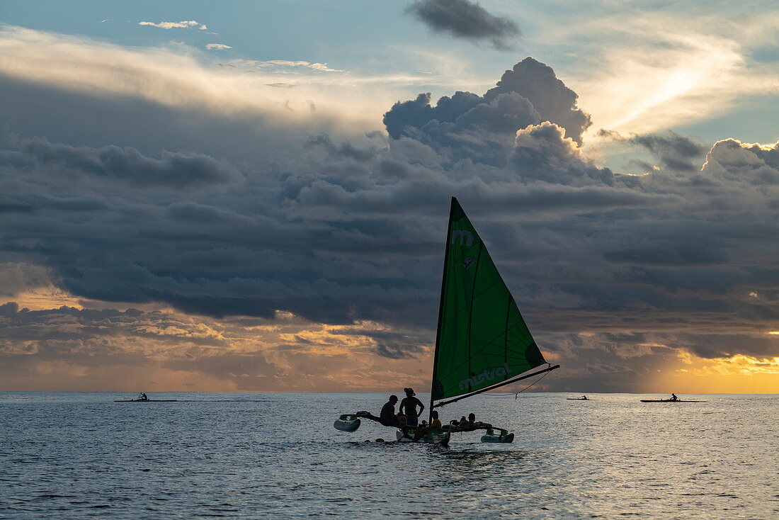 Menschen genießen Törn auf einem Auslegerkanu mit Segel bei Sonnenuntergang, nahe Papeete, Tahiti, Windward Islands, Französisch-Polynesien, Südpazifik