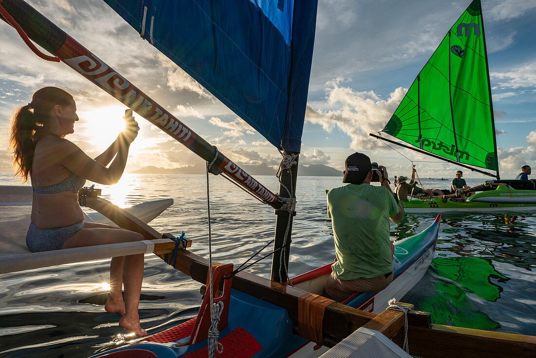 Menschen genießen Törn auf einem Auslegerkanu mit Segel bei Sonnenuntergang mit Blick auf die Insel Moorea, nahe Papeete, Tahiti, Windward Islands, Französisch-Polynesien, Südpazifik