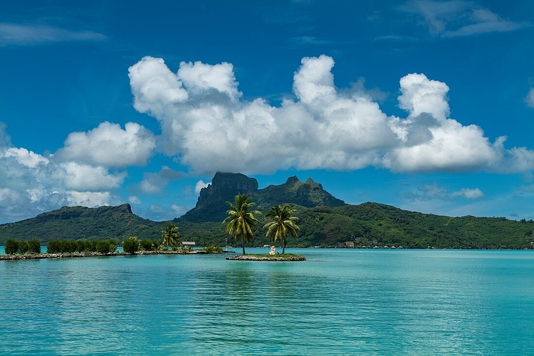 Two palm trees and a tiki sculpture on a small island at the entrance to Bora Bora Airport (BOB) harbor with Mount Otemanu in the distance, Bora Bora, Leeward Islands, French Polynesia, South Pacific