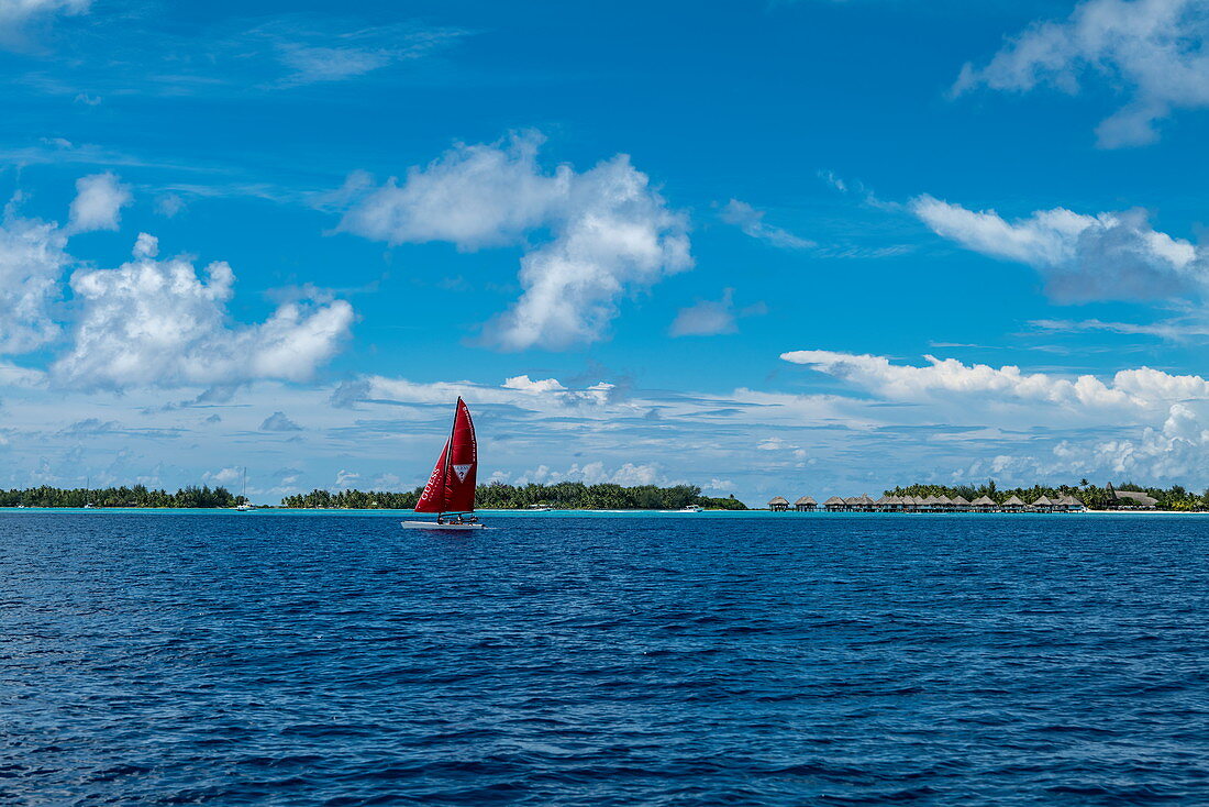 Auslegerkanu mit rotem Segel in der Lagune von Bora Bora mit Überwasserbungalows eines Resorts in der Ferne, Bora Bora, Leeward Islands, Französisch-Polynesien, Südpazifik