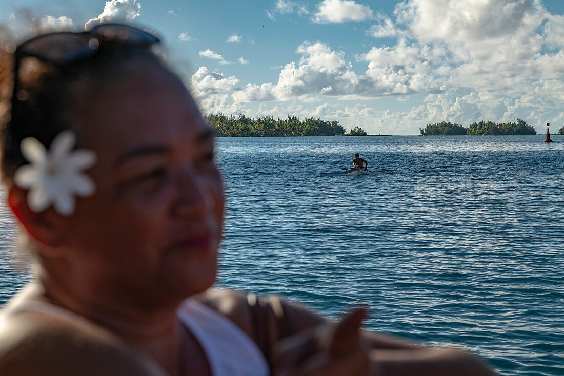 Outrigger canoe in the Bora Bora lagoon with woman in foreground carrying white flower behind her ear, Bora Bora, Leeward Islands, French Polynesia, South Pacific