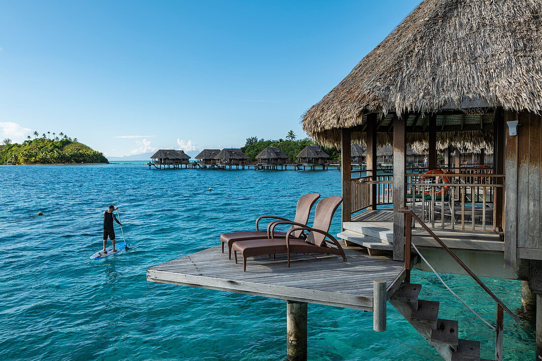 Mann auf SUP Stand Up Paddle Board passiert Überwasserbungalow des Sofitel Bora Bora Private Island Resort in der Lagune von Bora Bora, Bora Bora, Leeward Islands, Französisch-Polynesien, Südpazifik