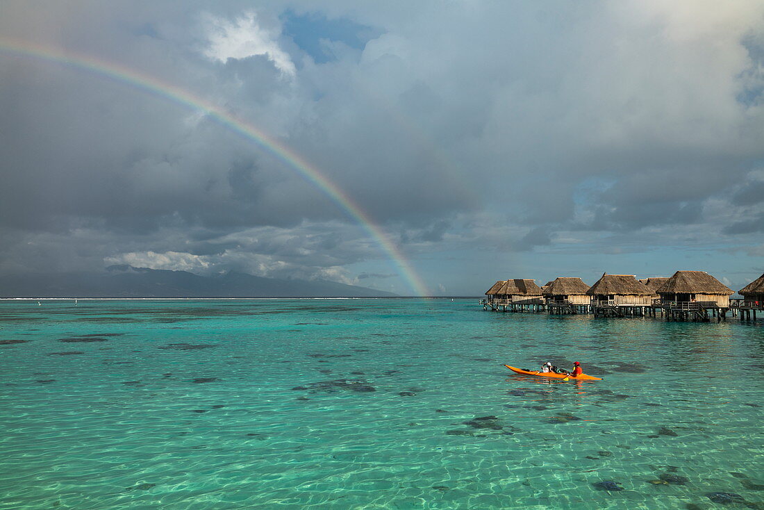 Menschen paddeln Kanu in der Lagune von Moorea mit Blick auf Regenbogen und Überwasserbungalows des Sofitel Ia Ora Beach Resort, Moorea, Windward Islands, Französisch-Polynesien, Südpazifik