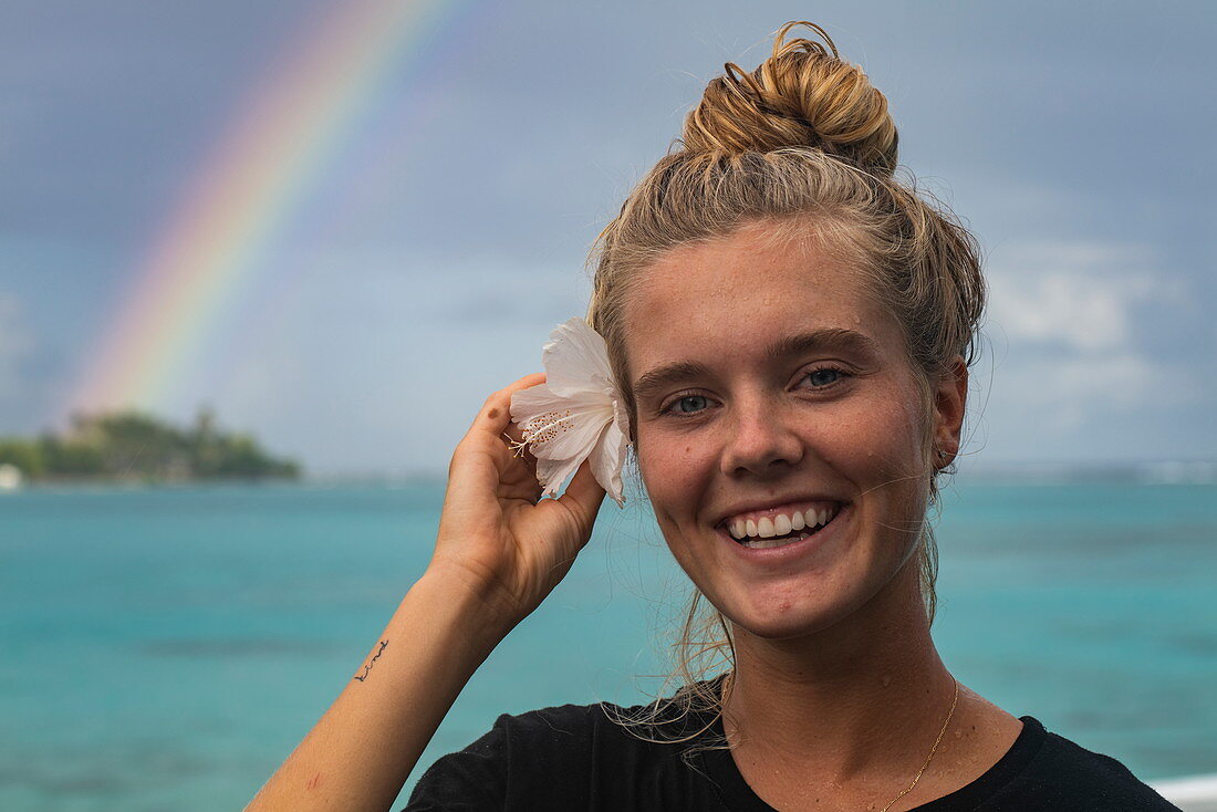Junge blonde Frau lächelt während sie eine Hibiskusblume hinter ihr Ohr steckt mit Regenbogen über der Lagune von Moorea dahinter, Moorea, Windward Islands, Französisch-Polynesien, Südpazifik