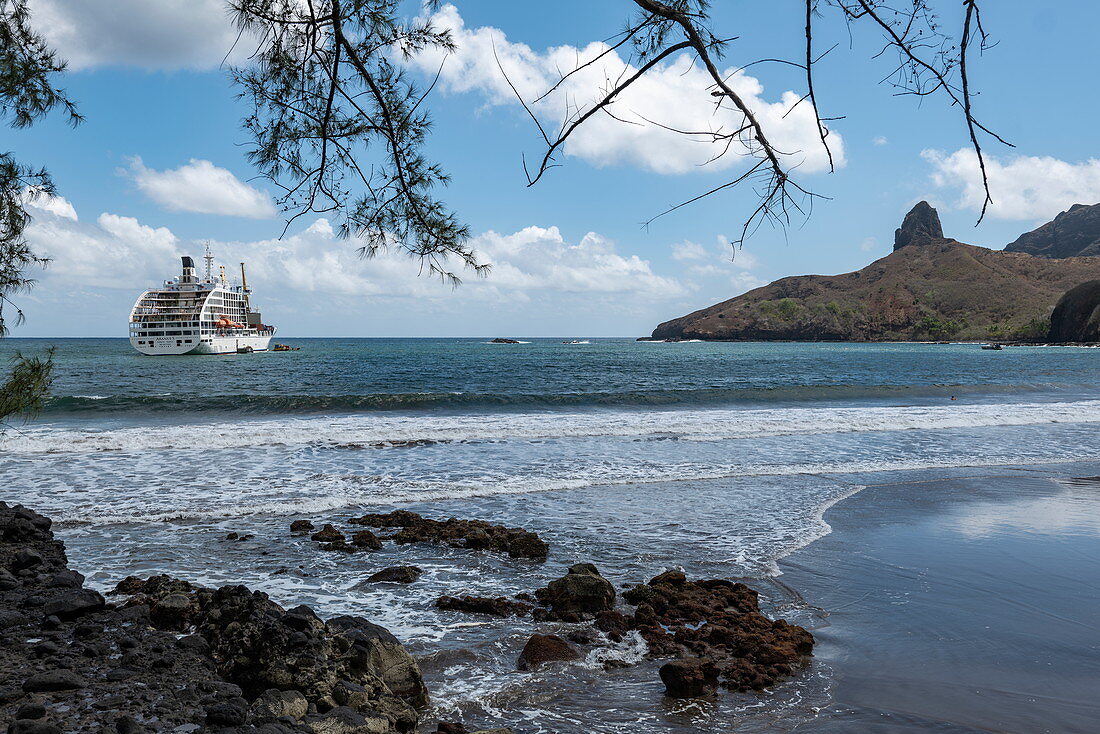 Felsen am schwarzen Sandstrand mit Passagierfrachtschiff Aranui 5 (Aranui Cruises) vor Anker in der Bucht, Puamau, Hiva Oa, Marquesas-Inseln, Französisch-Polynesien, Südpazifik