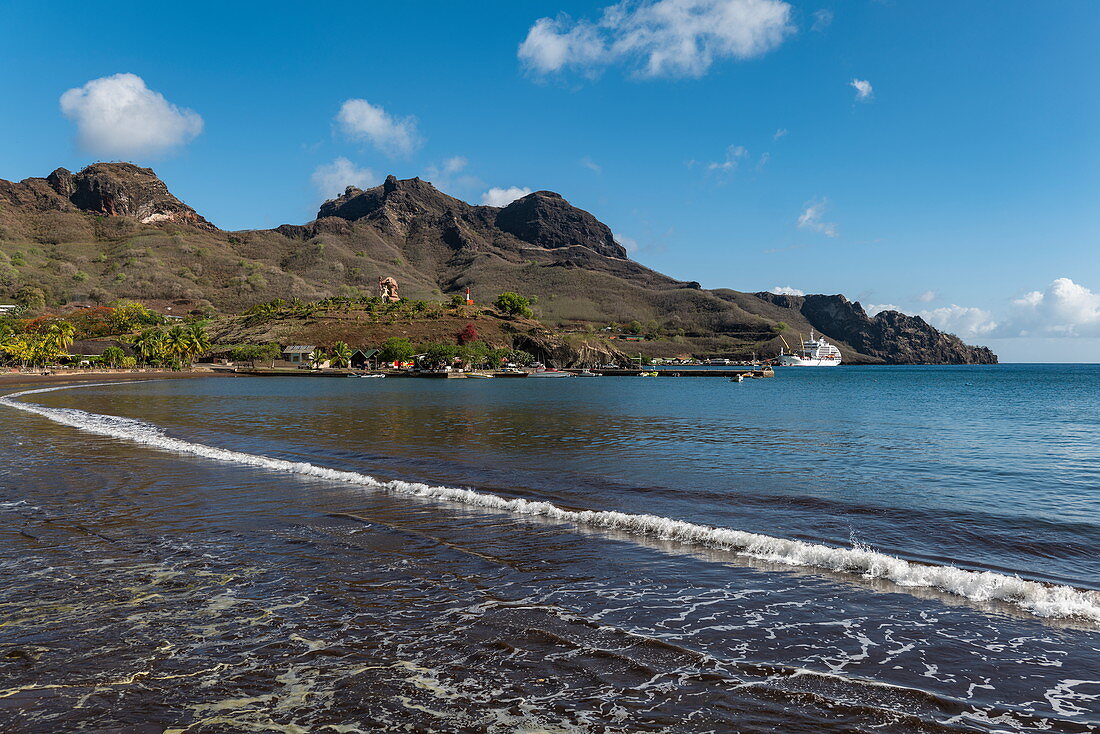 Schwarzer Sandstrand mit Passagierfrachtschiff Aranui 5 (Aranui Cruises) am Pier in der Ferne, Taiohae, Nuku Hiva, Marquesas-Inseln, Französisch-Polynesien, Südpazifik