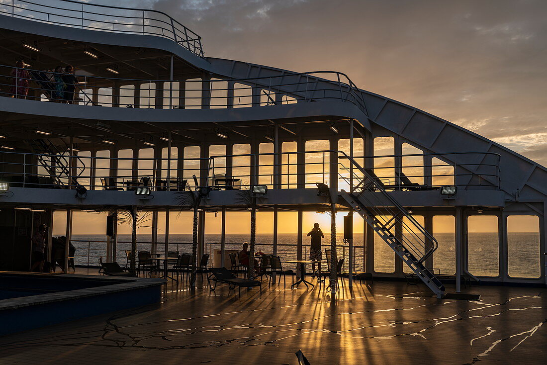 Silhouette von Menschen auf dem hinteren Sonnendeck von Passagierfrachtschiff Aranui 5  (Aranui Cruises) bei Sonnenuntergang, nahe Ua Pou, Marquesas-Inseln, Französisch-Polynesien, Südpazifik