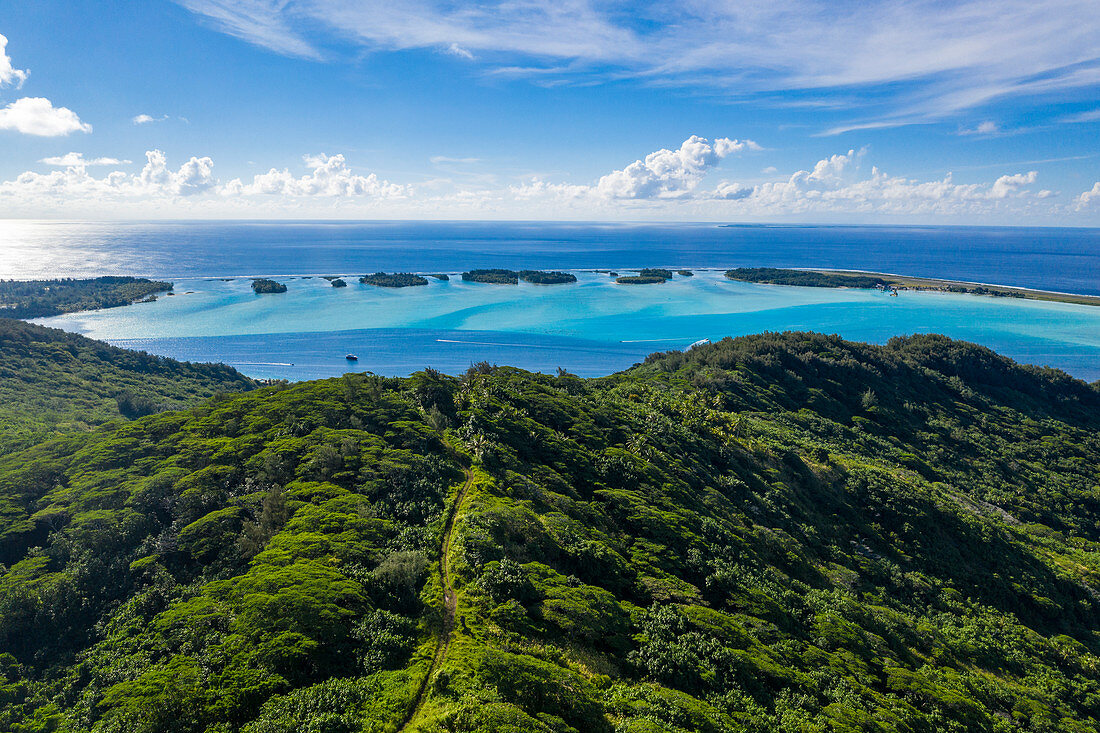 Luftaufnahme von unbefestigter Straße durch üppige Bergvegetation mit Motu Inselchen in der Lagune von Bora Bora in der Ferne, Bora Bora, Leeward Islands, Französisch-Polynesien, Südpazifik