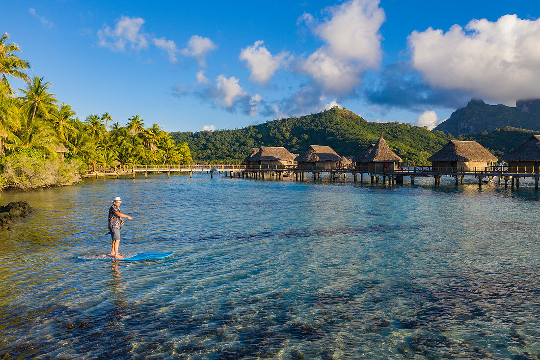 Luftaufnahme von Mann auf SUP Stand Up Paddle Board in der Lagune von Bora Bora mit Überwasserbungalows des Sofitel Bora Bora Private Island Resort dahinter, Vaitape, Bora Bora, Leeward Islands, Französisch-Polynesien, Südpazifik