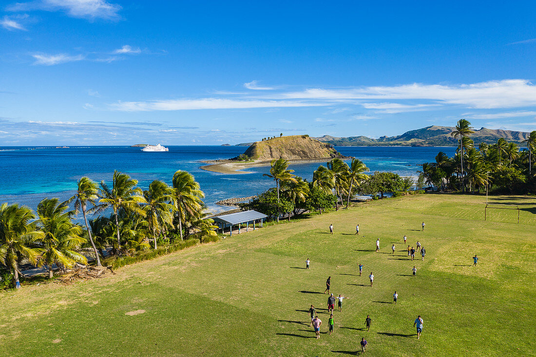 Luftaufnahme von Jungen die Rugby auf dem Feld der Dorfschule spielen mit Kreuzfahrtschiff MV Reef Endeavour (Captain Cook Cruises Fiji) in der Ferne, Nabukeru, Yasawa Island, Yasawa Group, Fidschi-Inseln, Südpazifik