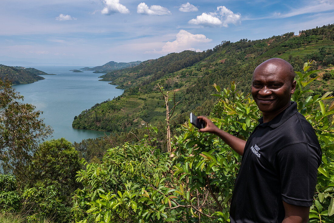 Mann mit Smartphone steht über Bucht am Kivu See, nahe Gitesi, Western Province, Ruanda, Afrika