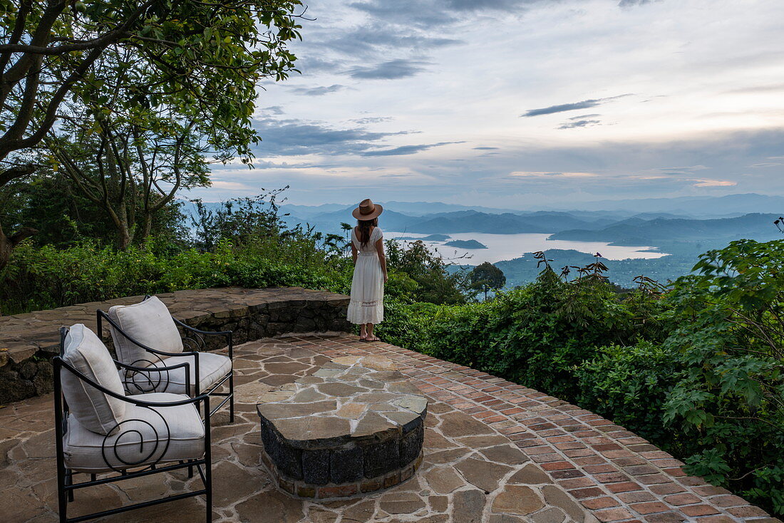 Young woman in summer dress and sun hat looks out over the Ruhondo Lake and the mountains from the terrace of the Virunga Lodge, near Kinyababa, Northern Province, Rwanda, Africa