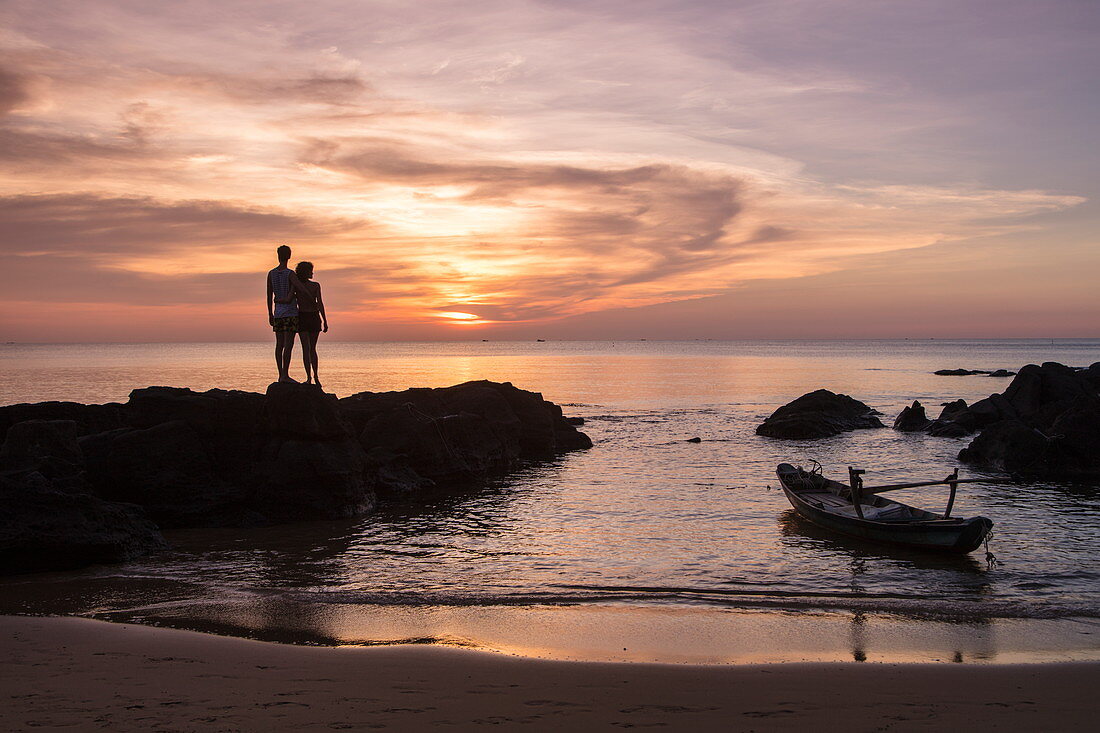 Silhouette von jungem Paar auf Felsen und Fischerboot am Ong Lang Beach bei Sonnenuntergang, Ong Lang, Insel Phu Quoc, Kien Giang, Vietnam, Asien
