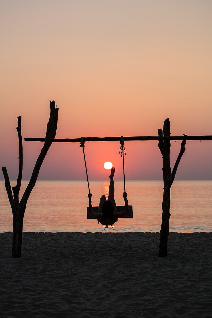 Silhouette von junger Frau auf Schaukel am Ong Lang Beach bei Sonnenuntergang die so tut als ob sie Sonne wie Fußball tritt, Ong Lang, Insel Phu Quoc, Kien Giang, Vietnam, Asien