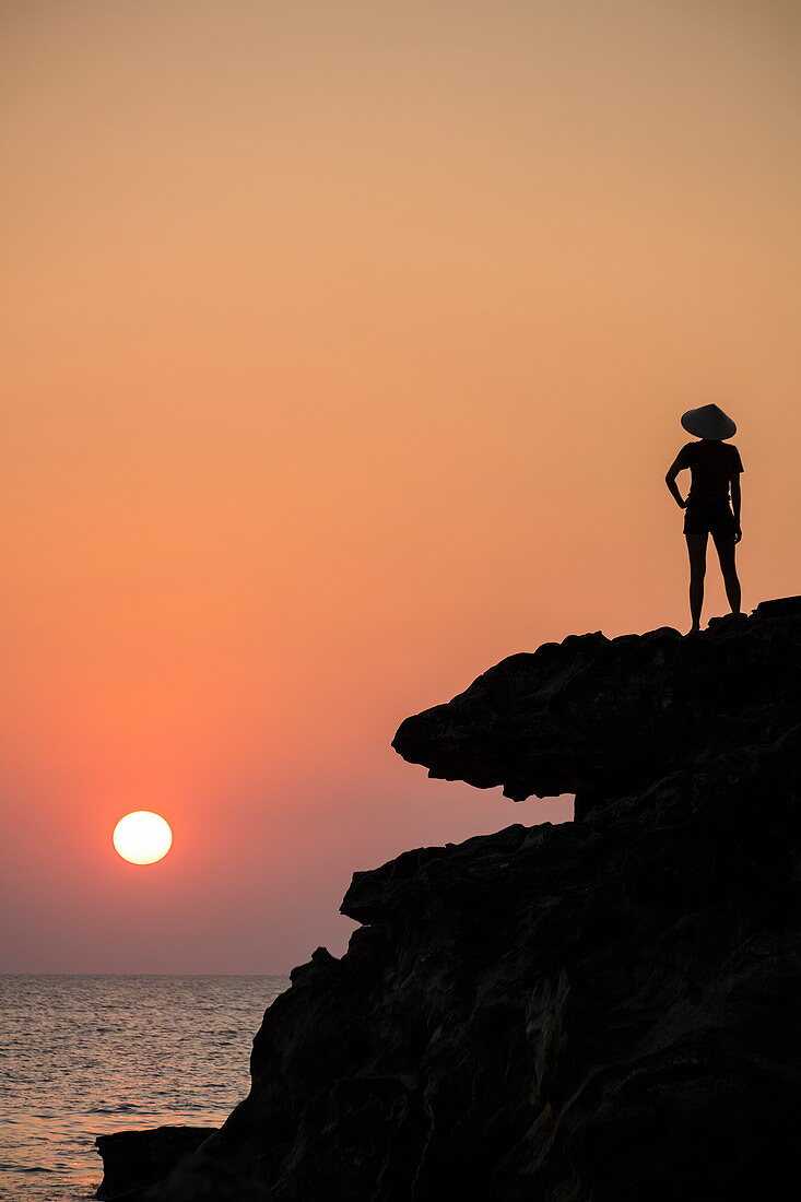 Silhouette von junger Frau die konischen Hut trägt und von Felsvorsprung neben Dinh Cao Schrein bei Sonnenuntergang auf Meer hinaus schaut, Duong Dong, Insel Phu Quoc, Kien Giang, Vietnam, Asien