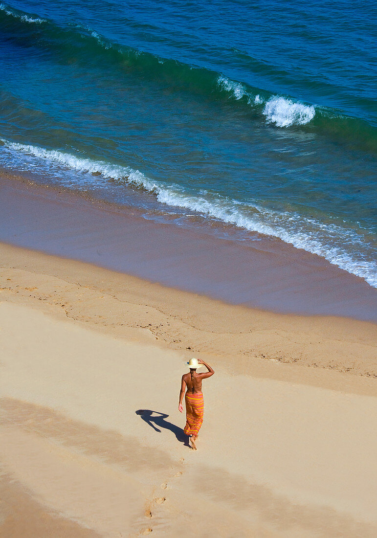 Frau in einem Sarong geht in goldfarbenem Sand auf das Wasser zu. Algarve, Portugal.