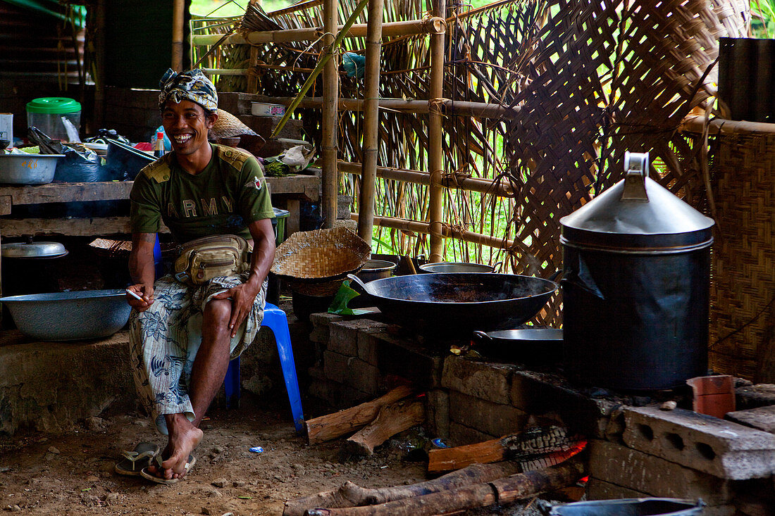 Ein balinesischer Koch, der in einer baufälligen Küche in einer Hütte sitzt, raucht und lacht. Bali, Indonesien, Asien