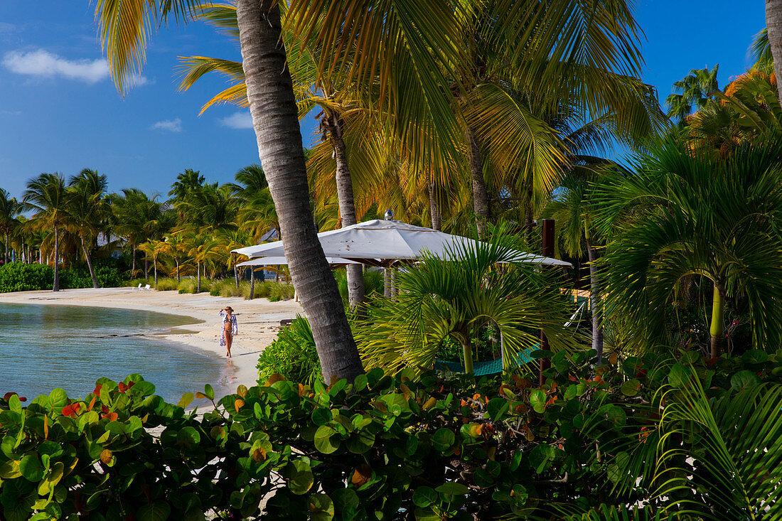 Frau, die an einem Strand entlang geht, der in einer sehr üppigen, tropischen Umgebung liegt, Sonnenschirme und Palmen, Karibik