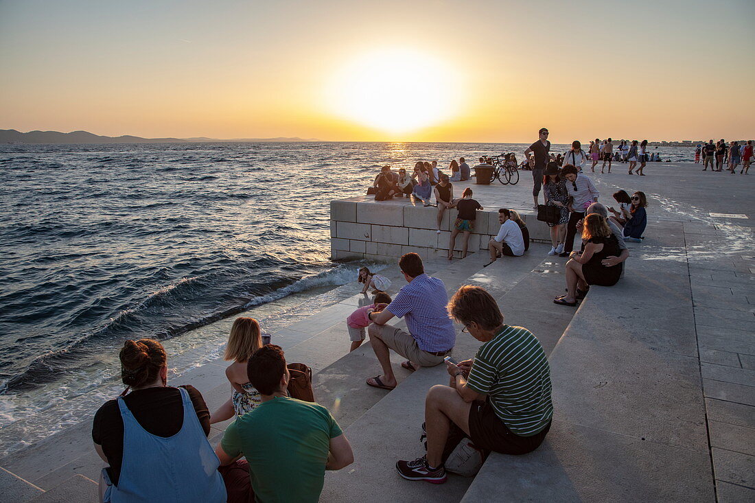 Menschen versammeln sich entlang der Strandpromenade um der Meeresorgel bei Sonnenuntergang zu lauschen, Zadar, Zadar, Kroatien, Europa