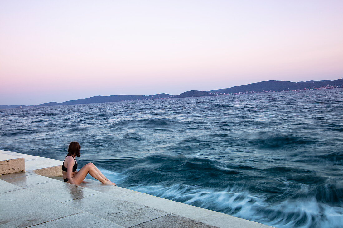 Junge Frau im Bikini sitzt auf Stufen der Strandpromenade bei Sonnenuntergang, Zadar, Zadar, Kroatien, Europa