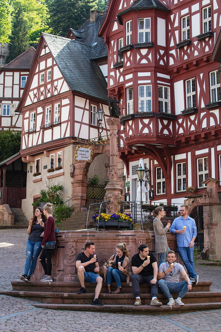 Young people enjoy ice cream while sitting around fountain with half-timbered buildings at the historic Schnatterloch market square, Miltenberg, Spessart-Mainland, Franconia, Bavaria, Germany, Europe