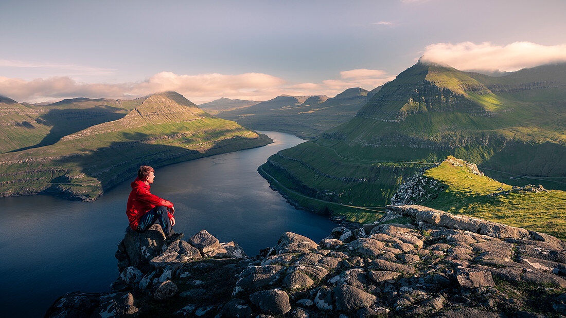 Mann sitzt am Hvithamar nahe dem Ort Gjogv auf der Färöer Insel Eysturoy mit Panoramablick in Fjord\n