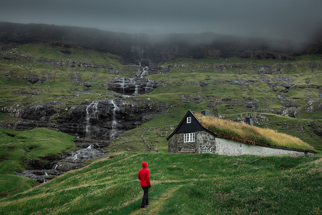 Man in red jacket in front of huts with grass roofs in the village of Saksun on Streymoy Island, Faroe Islands