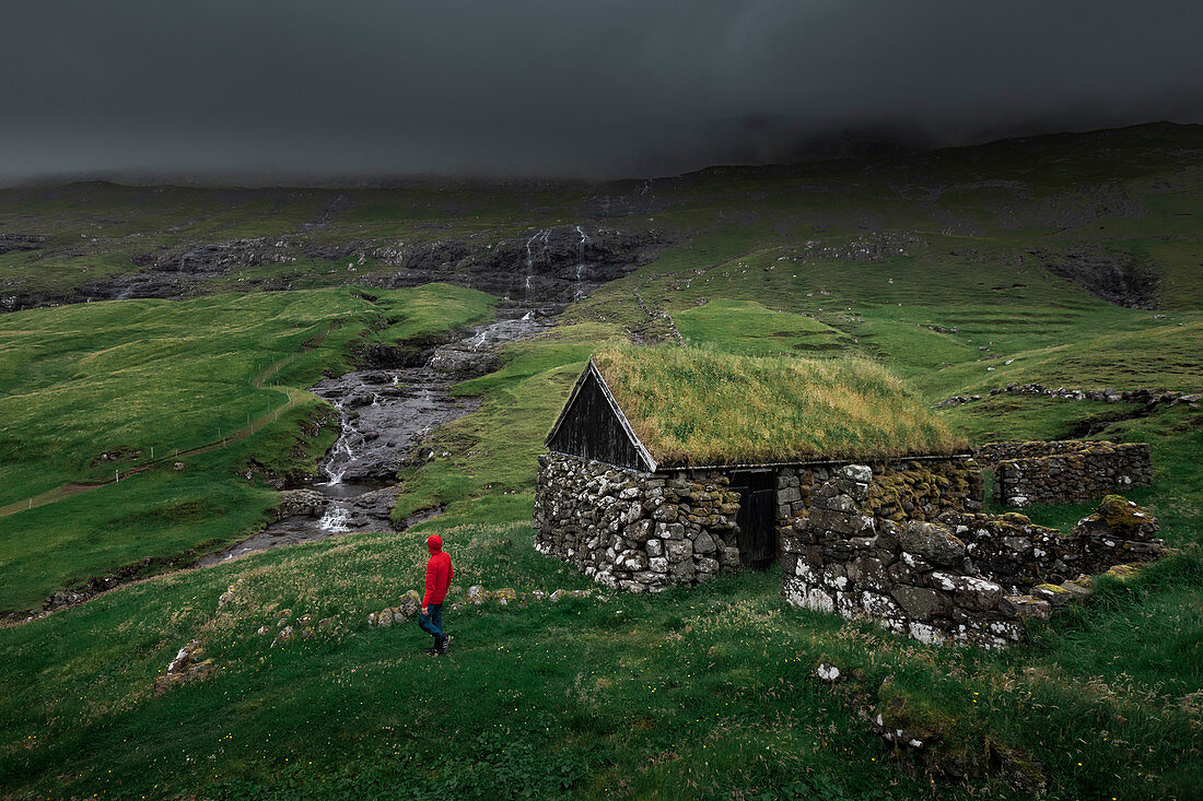 Mann in roter Jacke vor Hütten mit Grasdach im Dorf Saksun auf der Insel Streymoy, Färöer Inseln\n