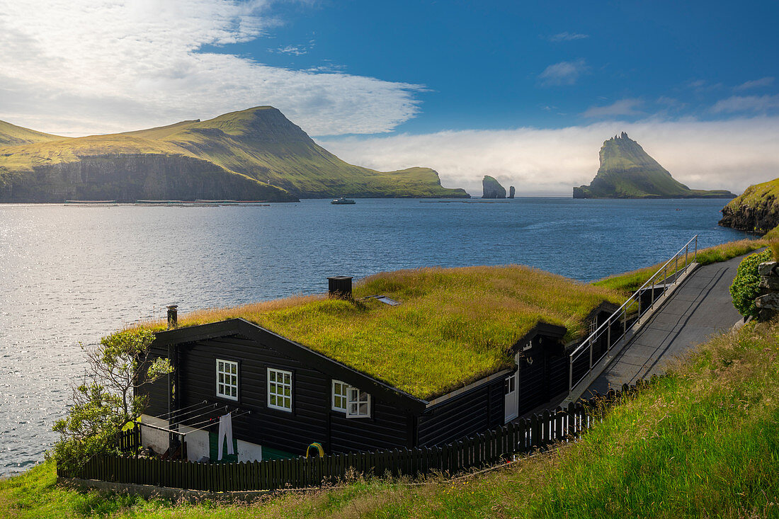 Wohnhaus mit Grasdach bei Sonne vor Felsformationen von Drangarnier auf Vagar, Färöer Inseln\n
