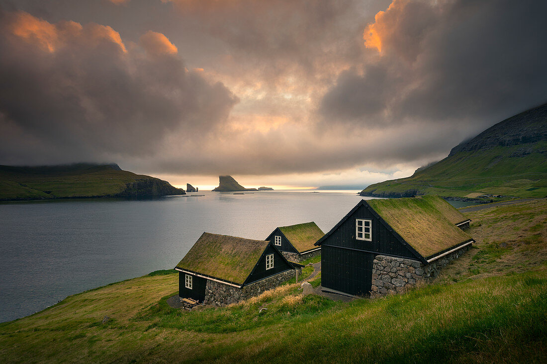 Häuser mit Grasdach im Sonnenuntergang vor Felsformationen von Drangarnier auf Vagar, Färöer Inseln\n
