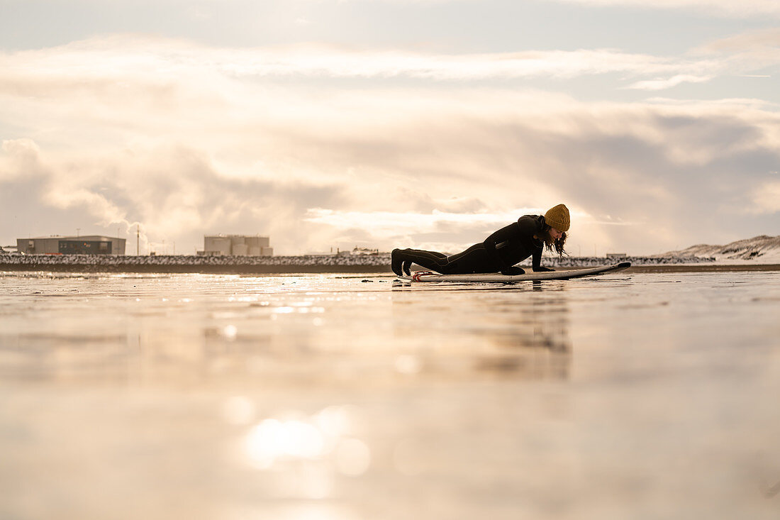 Silhouette einer Frau mit einem Surfbrett, das an einem Strand mit Industrieanlagen am fernen Horizont liegt.