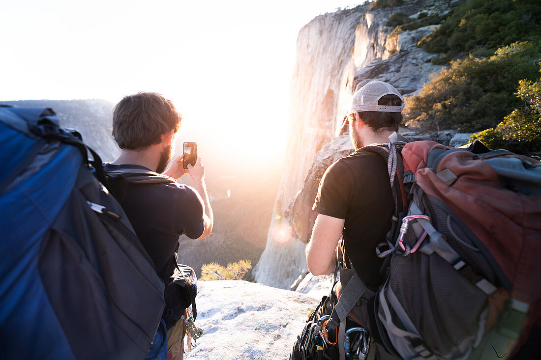 Rückansicht von zwei Männern, die Fotos von Felsen und einem Sonnenuntergang mit einem Mobiltelefon machen.
