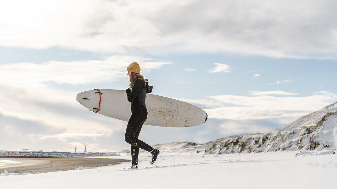 A woman wearing a wetsuit and holding a surfboard walking down a snowy beach and looking out to sea.