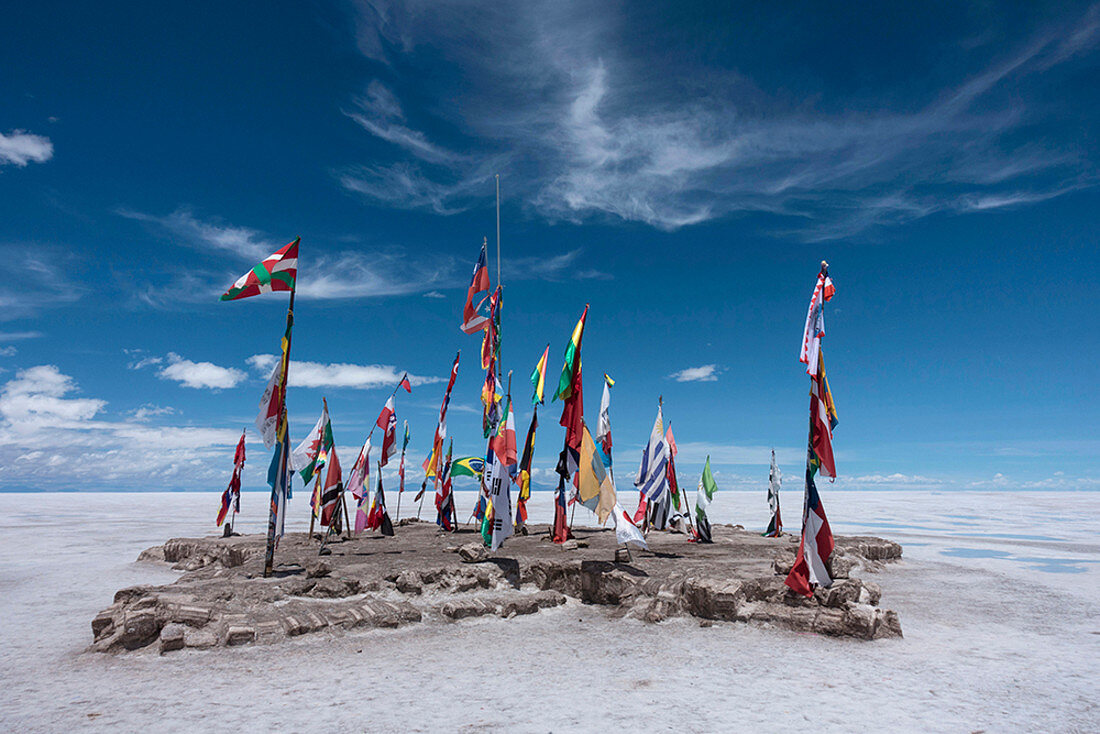 An outcrop of rocks on the salt flats with flags of many countries below a blue sky.
