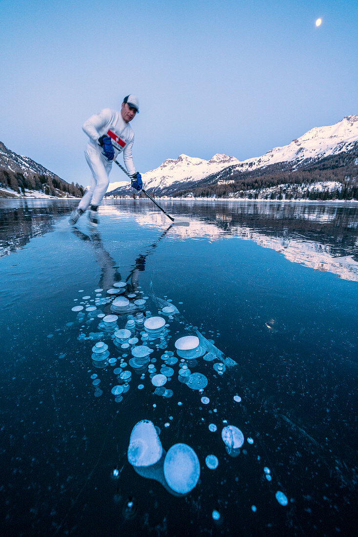 Ice hockey player man skating on Lake Sils covered in  ice bubbles at dusk, Engadine, Graubunden canton, Switzerland, Europe