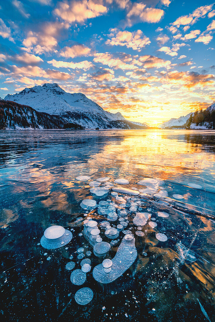 Wolken im brennenden Himmel bei Sonnenuntergang auf Piz Da La Margna und Eisblasen, die im Silssee, im Engadin, in Graubunden, in der Schweiz, in Europa eingeschlossen sind