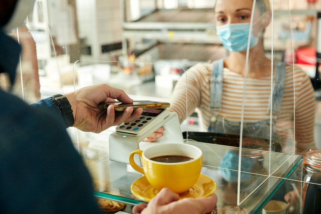 Frau mit Gesichtsmaske hinter Café-Theke mit Sicherheitsscheibe, die einem Kunden ein kontaktloses Zahlungsterminal anbietet