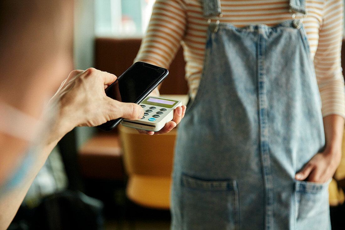 Frau mit Gesichtsmaske hinter Café-Theke hält kontaktloses Zahlungsgerät, während Kunde Handy verwendet, um Rechnung zu bezahlen