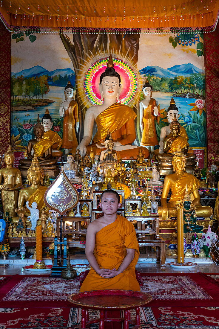 Junger Mönch posiert vor Buddha-Statue am Tempel Vat Chom Khao Manilat, Houayxay (Huay Xai), Provinz Bokeo, Laos, Asien