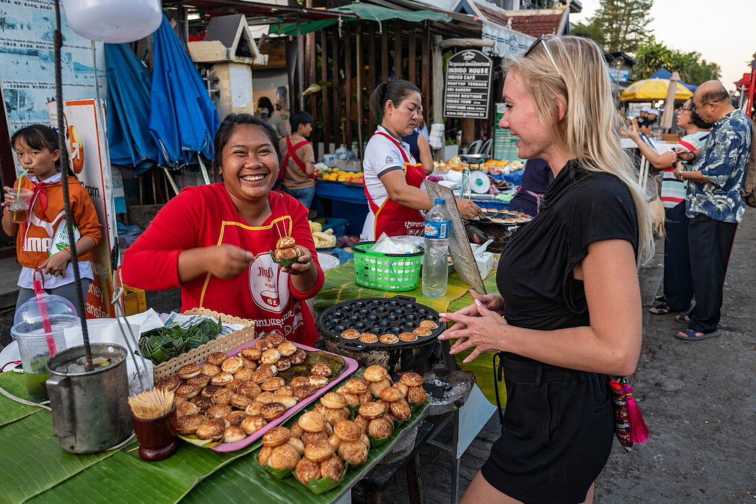 Junge blonde Frau lacht gemeinsam mit einer Verkäuferin von Kokosnusspfannkuchen an einem Verkaufsstand entlang der Sisavangvong Road (der Hauptstraße), Luang Prabang, Provinz Luang Prabang, Laos, Asien