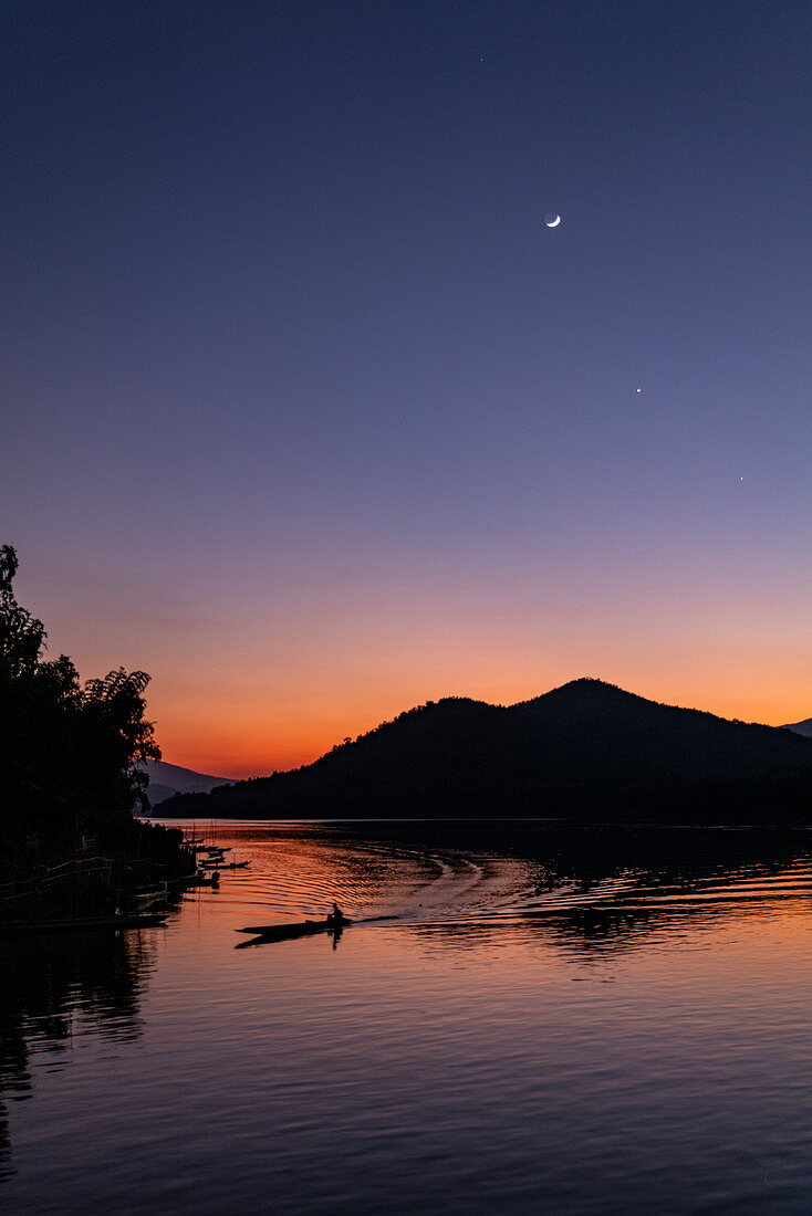 Silhouette von Longtail-Boot auf Fluss Mekong und Berge bei Sonnenuntergang mit Mondsichel und Venus, Luang Prabang, Provinz Luang Prabang, Laos, Asien