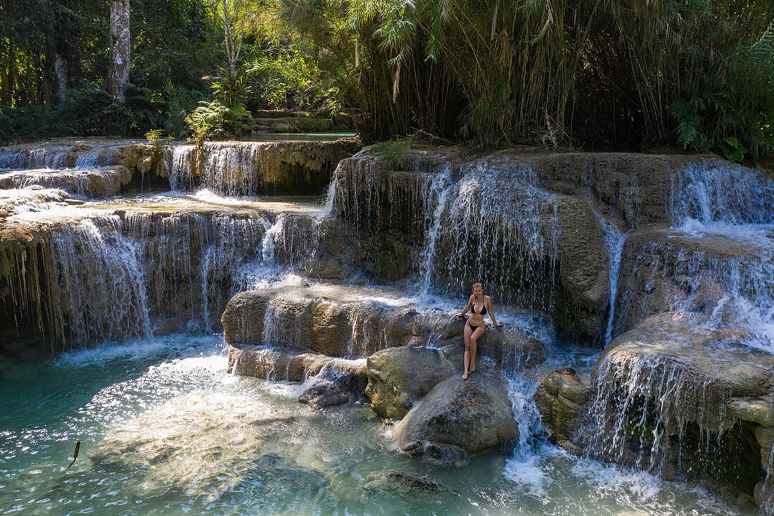 Aerial view of a beautiful young blonde woman on a rock in the natural pools of the magnificent Kuang Si Falls, Kuang Si, Luang Prabang Province, Laos, Asia